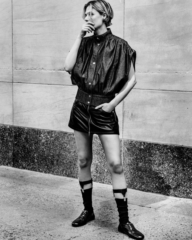 Louisa Jacobson standing outdoors in a sleeveless dark top, zip mini skirt, and garter socks in a black and white fashion portrait.