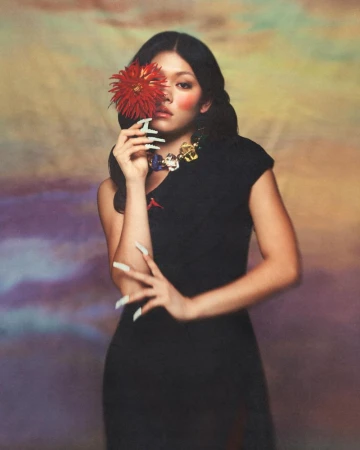 Momona Tamada in a black dress holding a red flower over one eye against a painterly gradient backdrop.