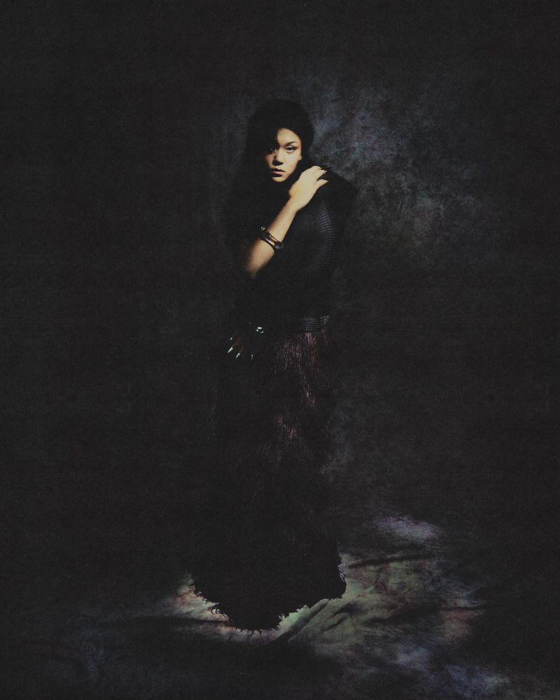 Actress Momona Tamada posing in a dark textured gown under low-key lighting against a mottled dark studio backdrop.