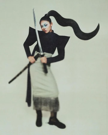 Momona Tamada poses with a katana wearing a black structured top with sharp shoulder wings and an arched ponytail.