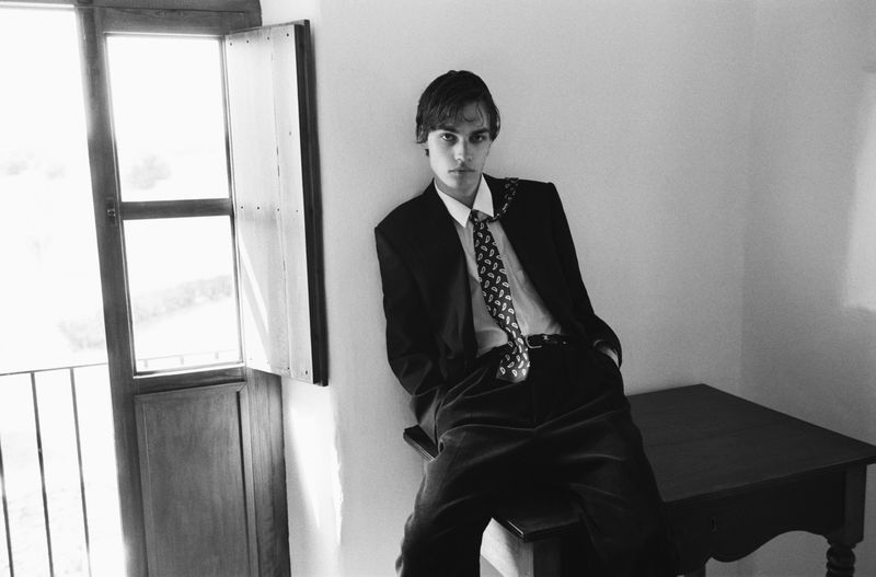 Model Bastian Bergstrøm in a dark suit, white shirt, and patterned tie sitting on a table by an open window in black and white.