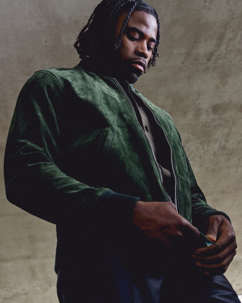 Black man wearing a dark green suede bomber jacket against a textured concrete wall.