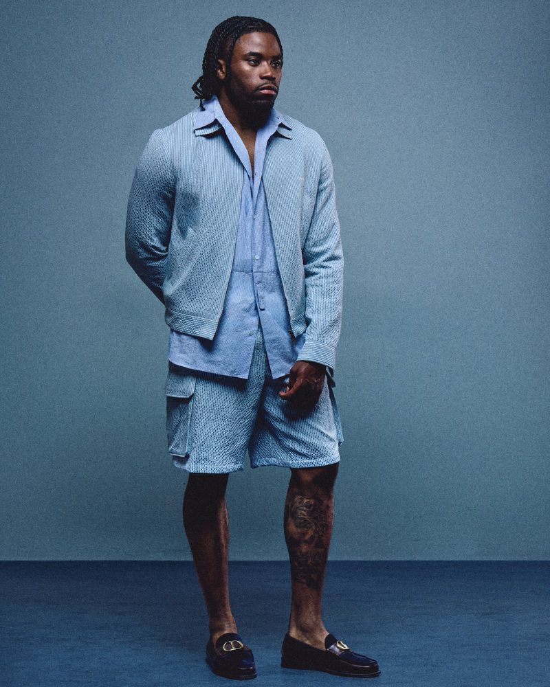 Full-body portrait of Kenny Moore II wearing a light blue textured jacket, matching shirt, and shorts against a solid grey-blue background.