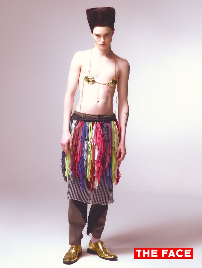 Model Malte Carlsson posing shirtless with tall structured hair, gold chain jewelry, colorful fringe skirt, and metallic gold shoes.
