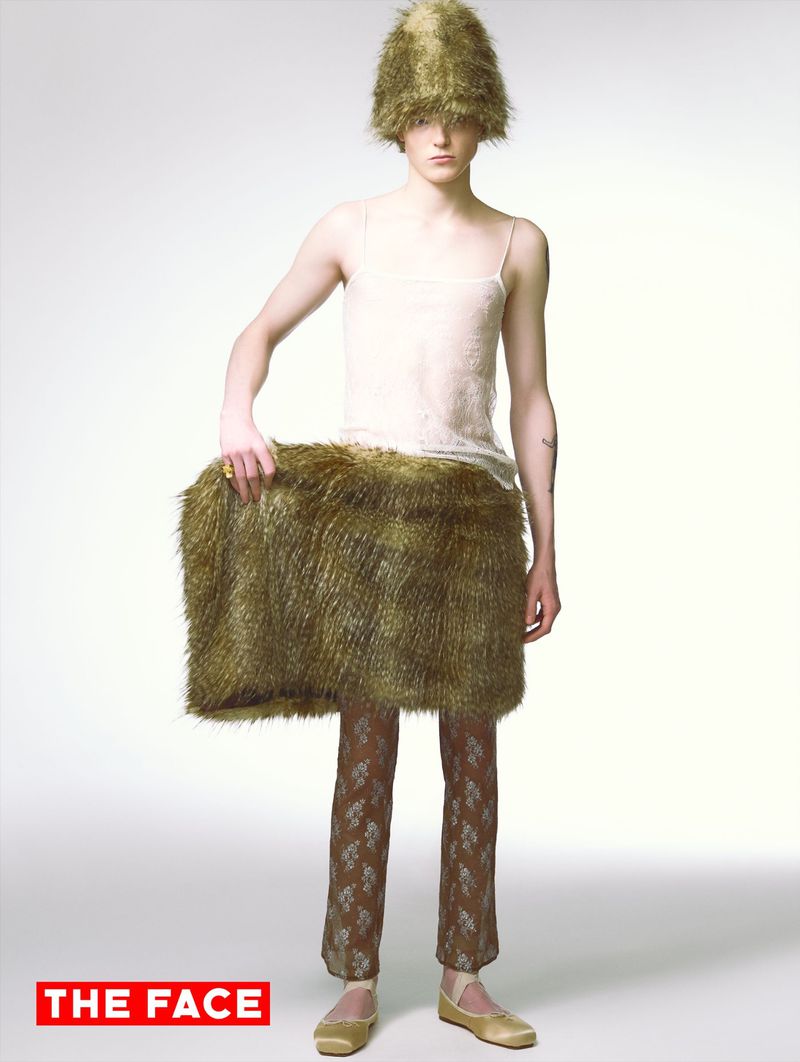 Model Malte Carlsson wears a fuzzy green hat, white camisole, wide textured green panel, patterned trousers, and flats on white background.