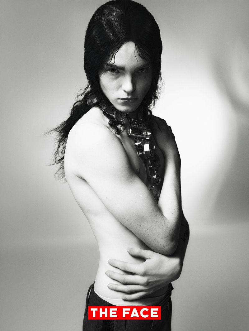 Black and white editorial portrait of a shirtless male model with long dark hair wearing a sculpted neckpiece against a studio backdrop.