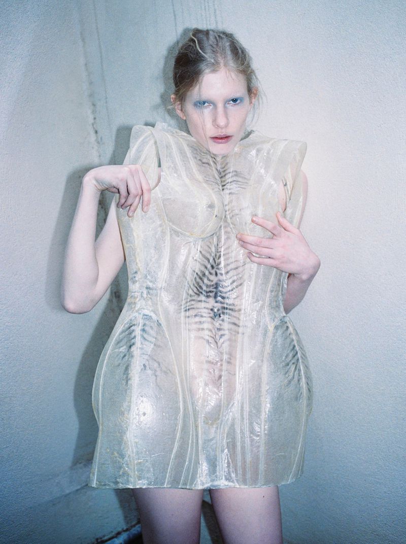 Model Victoria Volt in a structured translucent corset dress posing against a pale textured wall in direct flash lighting.