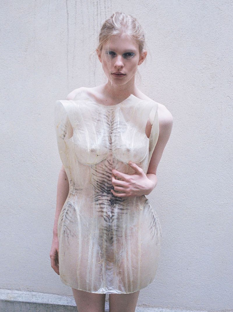 Model Victoria Volt wearing a sheer sculptural molded mini dress with skeletal motifs against a textured pale wall.