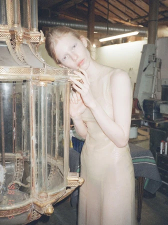 Model wearing a sheer beige dress leaning against a large ornate glass fixture in an industrial workshop.