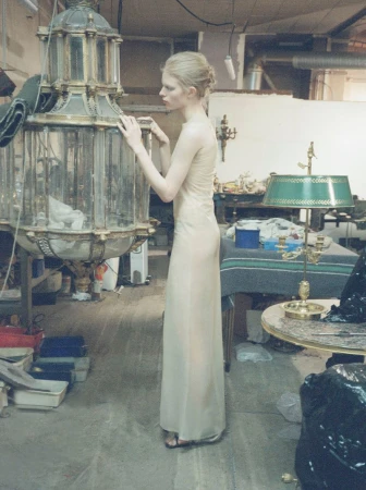 Model Victoria Volt wearing a long sheer cream dress standing beside an antique glass lantern in a workshop setting.