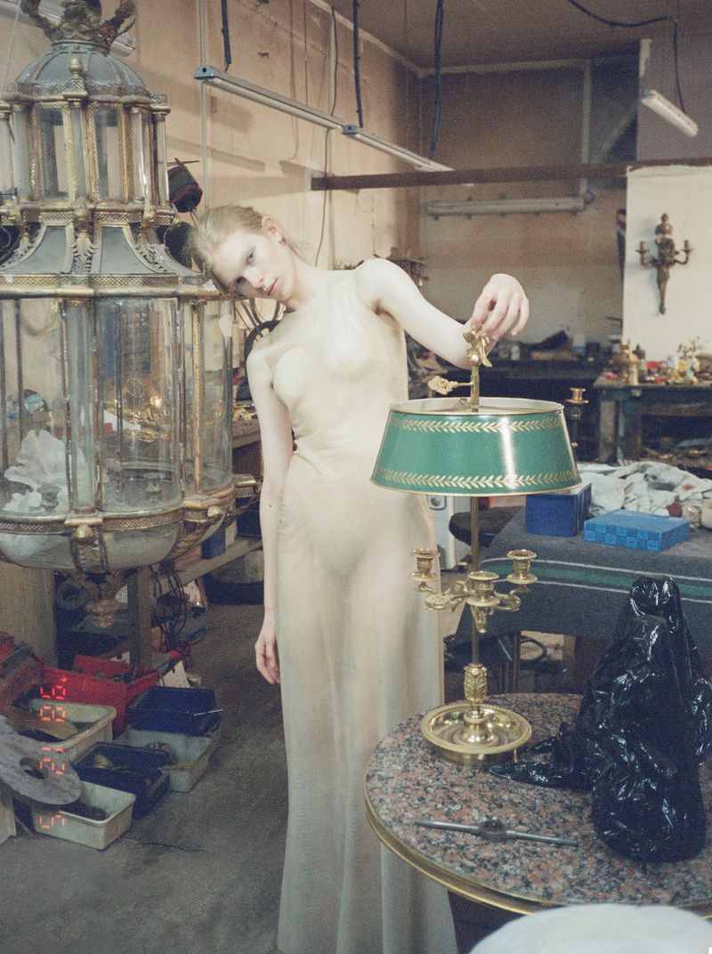 Model wearing a sheer long dress standing in a workshop interior next to a vintage brass lamp and large lantern