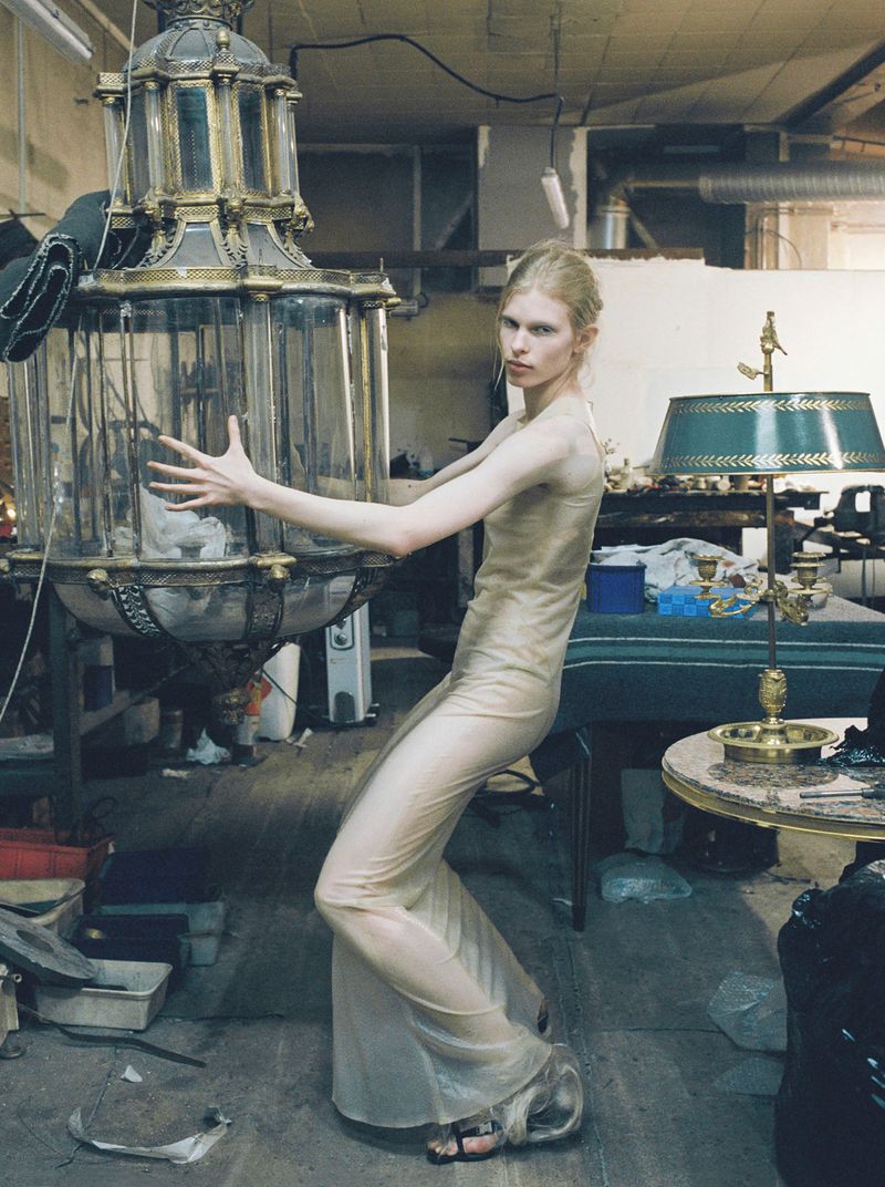 Model Victoria Volt in a sheer beige gown posing dynamically beside a large antique lantern chandelier in a workshop setting.