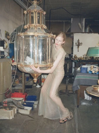 Model Victoria Volt posing in a sheer gown while holding a large antique glass lantern in an industrial workshop.