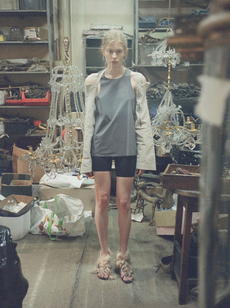 Model Victoria Volt in a grey top, black shorts, and detached beige sleeves standing among crystal chandeliers in a workshop.