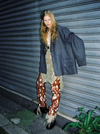 Model with long blonde hair wearing a dark grey oversized blazer and cow print patterned trousers leaning against a metal shutter door.