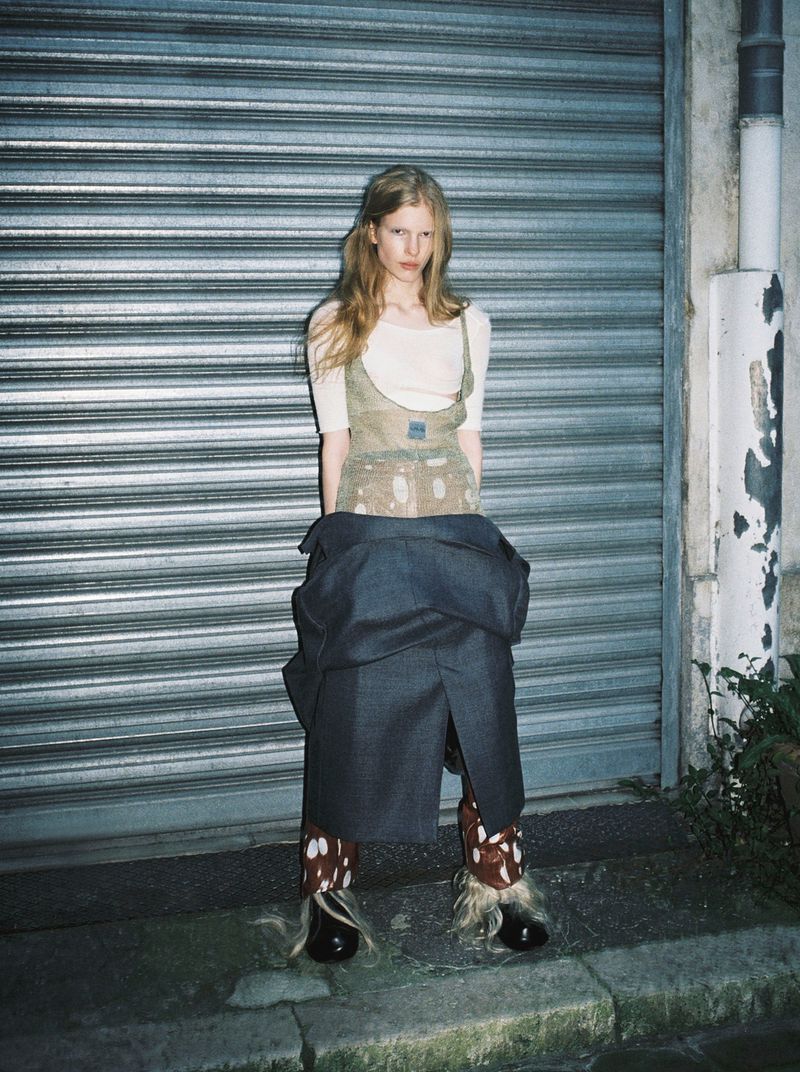Model Victoria Volt poses in a layered avant-garde outfit with a charcoal coat tied around her waist before a metal shutter.