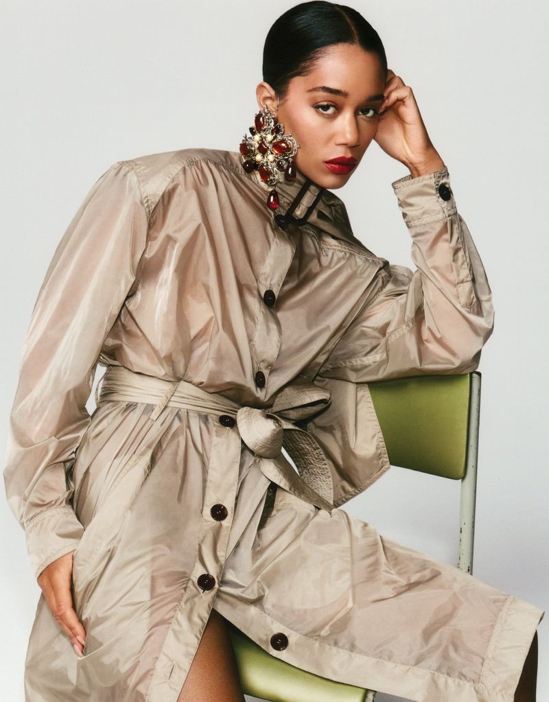 Laura Harrier wearing a beige belted coat and ornate crystal earrings while seated on a green chair