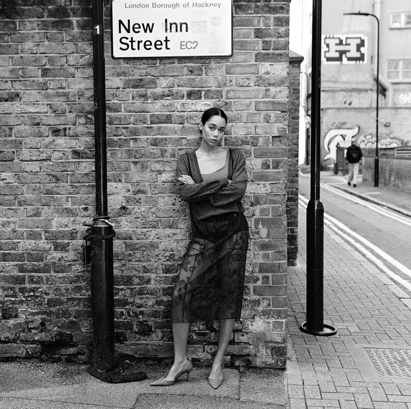 Laura Harrier leaning against a brick wall beneath a New Inn Street sign on a London street.