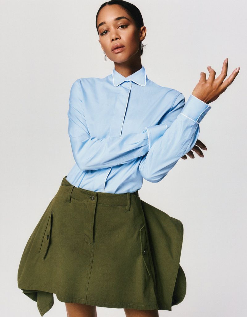 Laura Harrier posing in a light blue collared shirt and olive green structural mini skirt against a white studio background.