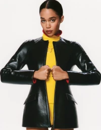 Actor Laura Harrier poses in a black leather jacket and yellow turtleneck against a white studio background.
