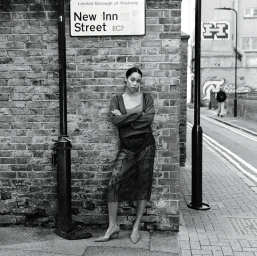 Laura Harrier leaning against a brick wall beneath a New Inn Street sign on a London street.