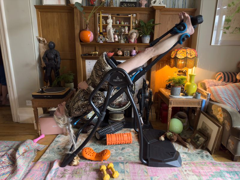 Woman in inversion table inside a residential living room with wood cabinet, vintage furniture, and patterned area rug.  