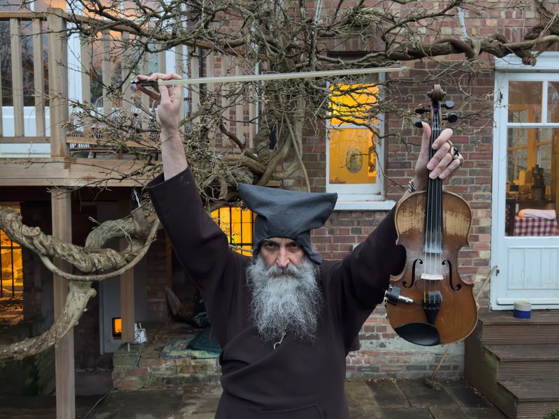 Adult male with long beard stands outside, holding violin and bow aloft with weathered brick building and tree branches in background.