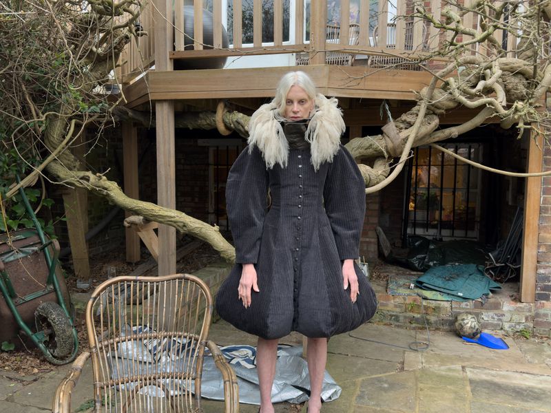 An adult woman with pale skin and grey hair stands outdoors in a garden wearing a puffy black dress with a white fur collar.