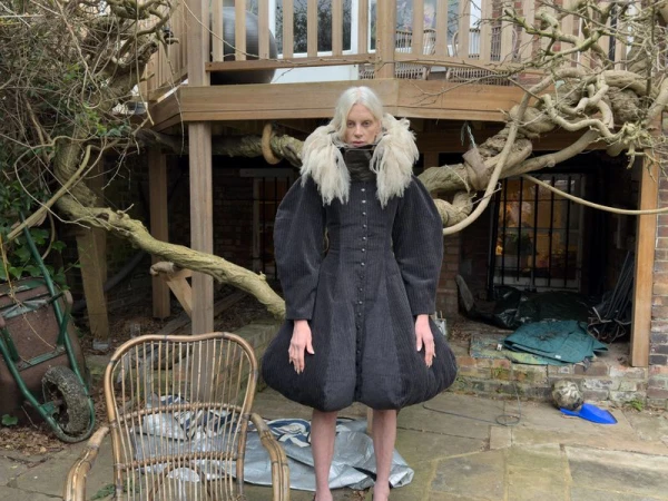 An adult woman with pale skin and grey hair stands outdoors in a garden wearing a puffy black dress with a white fur collar.