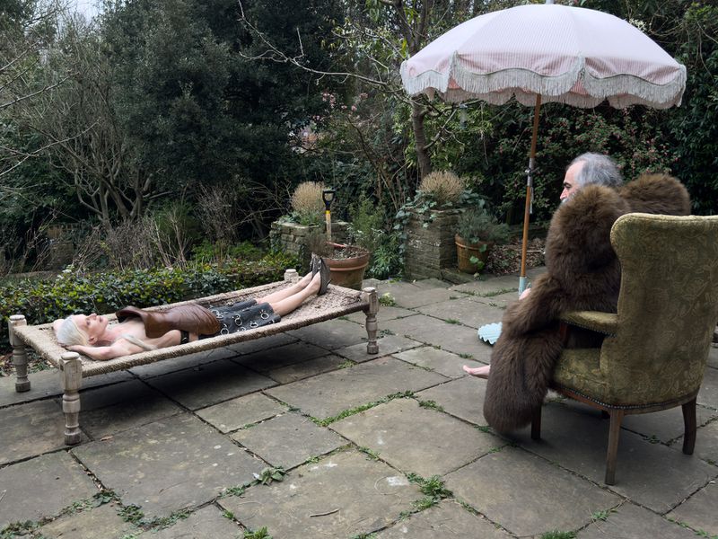 Outdoor scene featuring an adult male in a brown fur coat sitting in a green chair, with another adult relaxing on a woven daybed.