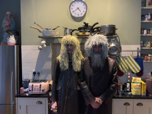Two people in a kitchen wearing shaggy wigs and long coats. Kitchen interior includes appliances, cabinets, and a religious statue.