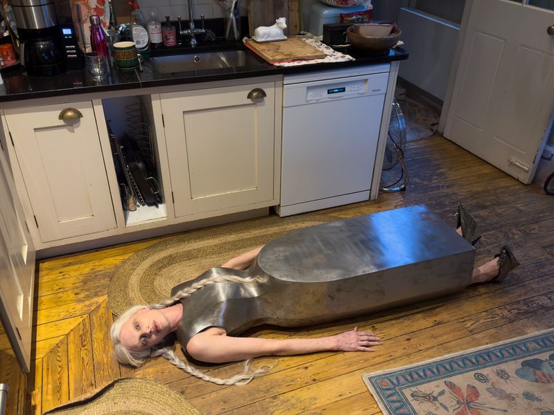 Unusual kitchen image: Woman in metallic garment lays on wooden floor near cabinets, embodying a blend of fashion and interior elements.
