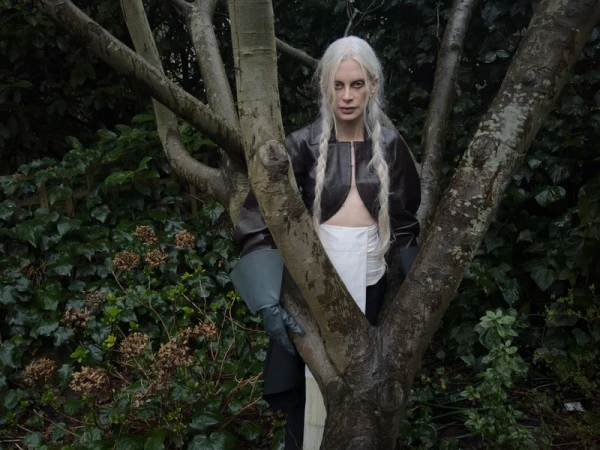 Adult woman with braided white hair stands in a garden in a brown leather jacket and white skirt beside a tree trunk.