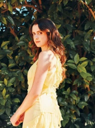 Young woman with long brown hair wearing a yellow ruffled dress outdoors in a leafy garden.