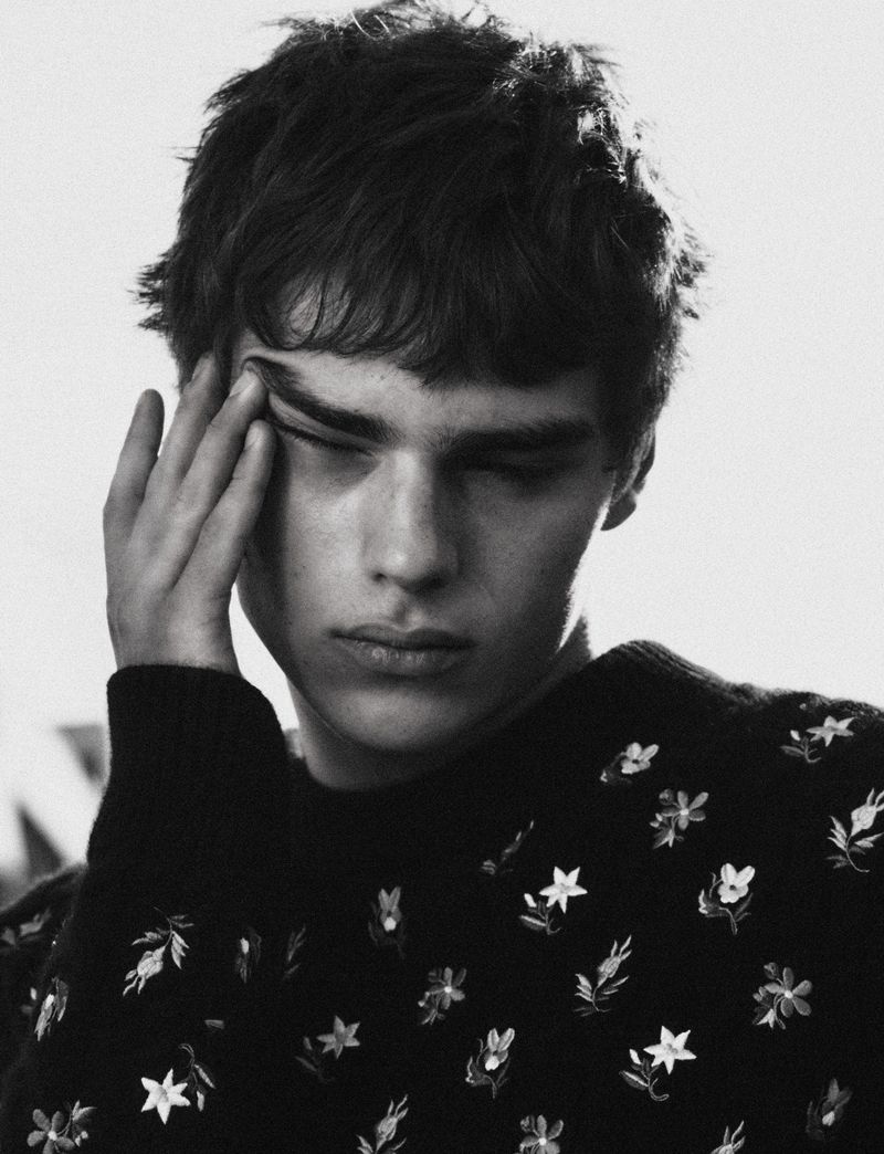 Young male model wearing a dark embroidered floral sweater with hand touching temple against plain background