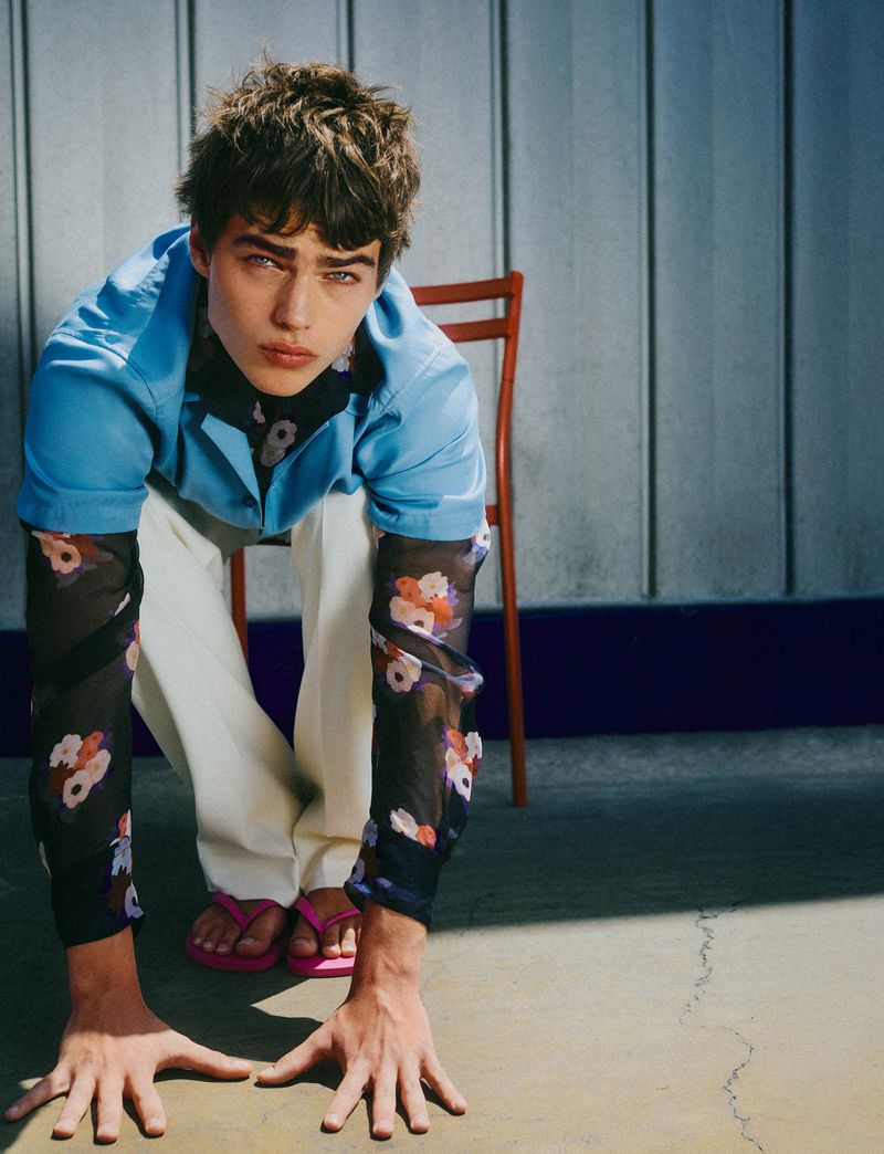 Model Jon Payeras crouching forward with hands flat on the concrete wearing a blue shirt, floral sheer sleeves, and white trousers.