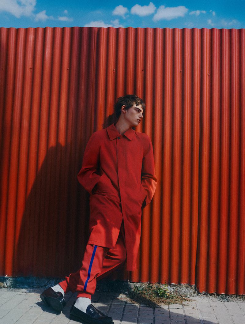 Man wearing a red coat and red trousers standing against a corrugated red wall under a blue sky.