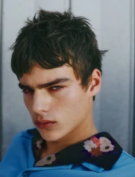 Close-up portrait of model Jon Payeras wearing a bright blue shirt with a floral collar against a grey background.
