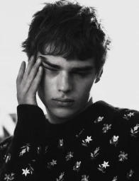 Young male model wearing a dark embroidered floral sweater with hand touching temple against plain background