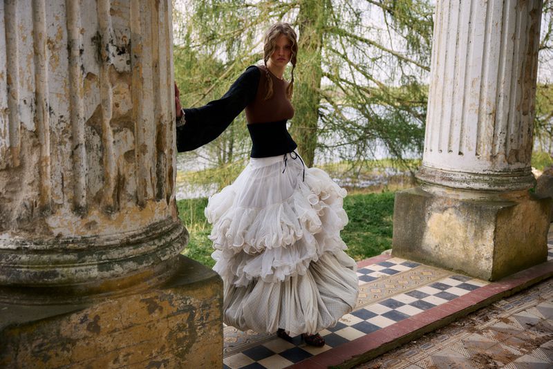 Model Batura Aliaksandra in a tiered white ruffle skirt and corset posing between weathered stone columns outdoors.