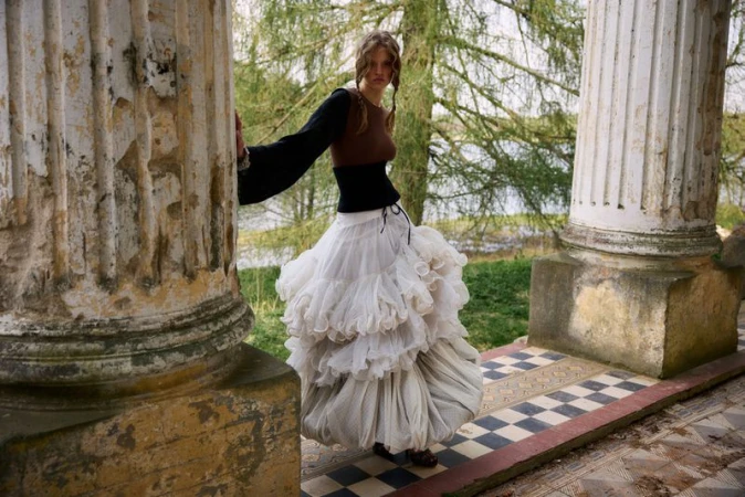 Model Batura Aliaksandra in a tiered white ruffle skirt and corset posing between weathered stone columns outdoors.