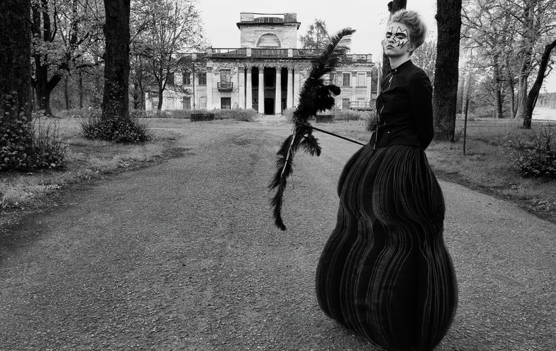 Model in a sculptural black skirt holding a feathered staff on a path leading to a neoclassical manor in black and white.