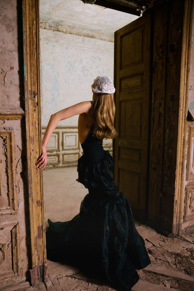 Model seen from behind wearing a black backless gown and white headpiece in the doorway of a weathered room.