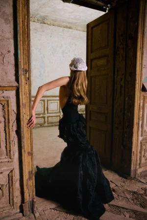 Model seen from behind wearing a black backless gown and white headpiece in the doorway of a weathered room.