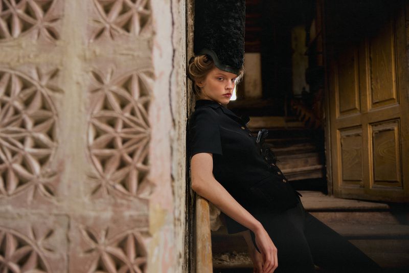 Model wearing a black tailored outfit and tall textured headpiece leaning against an ornate carved stone wall inside an aged castle interior.