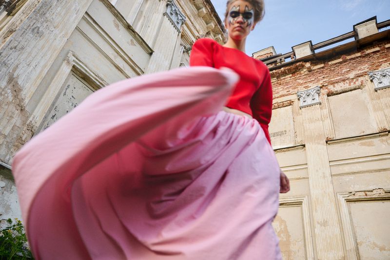 Low-angle shot of a model in a red top and billowing pink skirt posing in front of a weathered classical building with face paint.