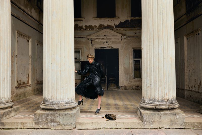Model Batura Aliaksandra wearing a voluminous black dress posing between stone columns of a neoclassical building with a cat nearby.