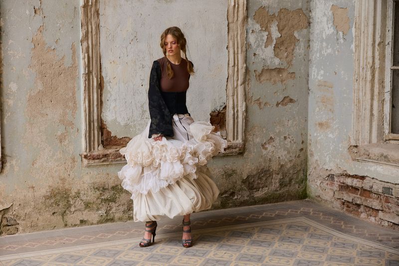 Model Batura Aliaksandra posing in a voluminous white ruffled skirt and brown bodice against distressed stone walls.