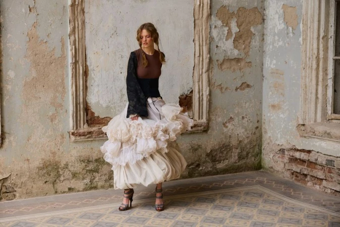 Model Batura Aliaksandra posing in a voluminous white ruffled skirt and brown bodice against distressed stone walls.