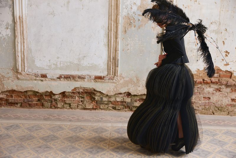Model wearing a black sculptural pleated skirt and black feather accessory standing against a weathered wall with exposed brick.